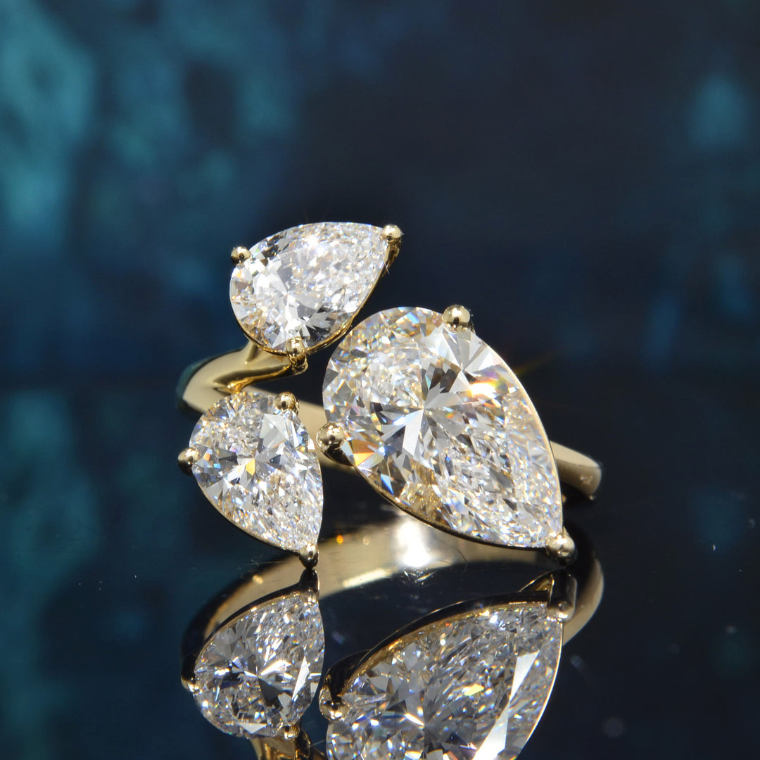 A gold ring featuring three pear-cut diamonds in prong settings, displayed on a reflective dark blue surface.