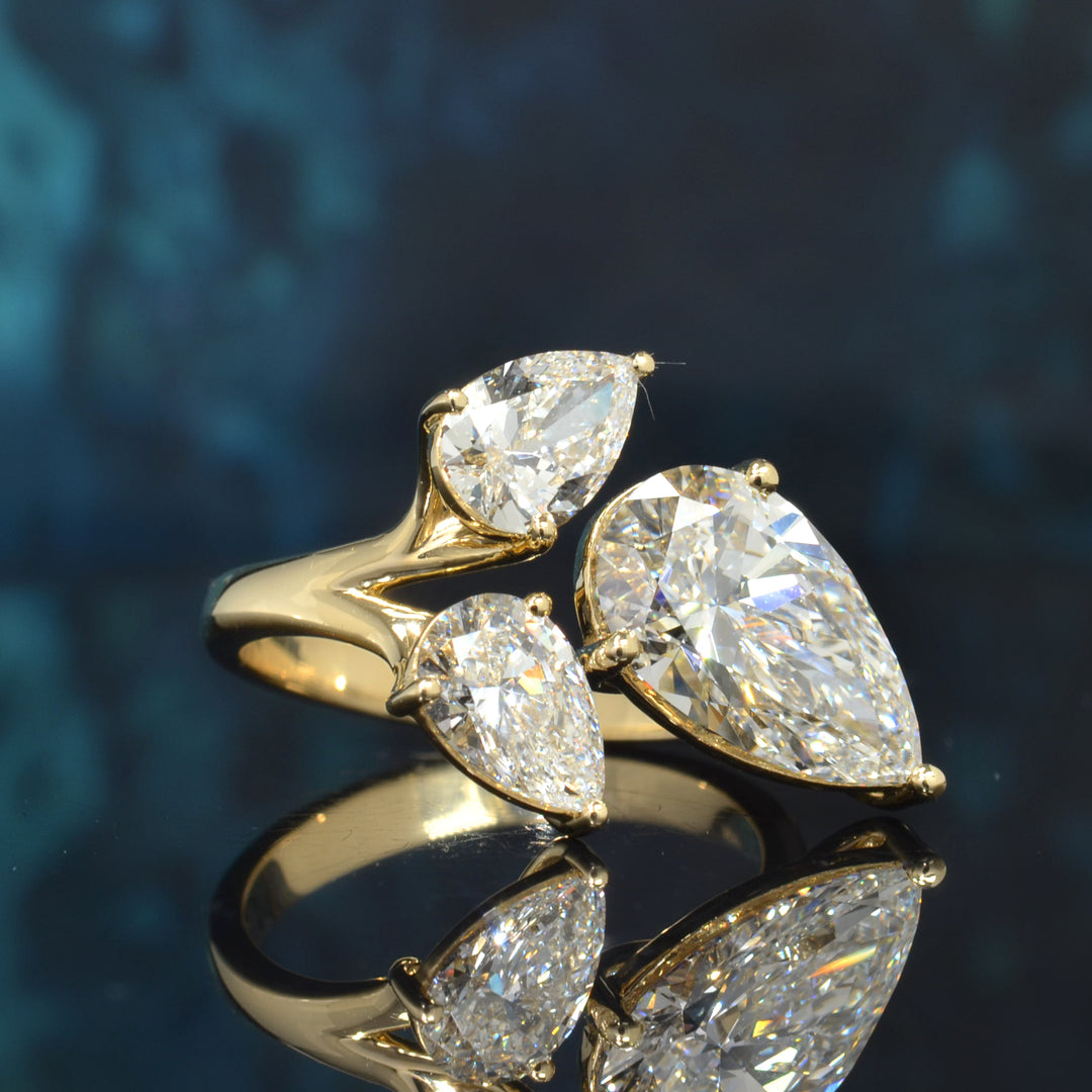 A gold ring featuring three pear-cut clear gemstones in a cluster setting, displayed on a reflective dark blue surface.