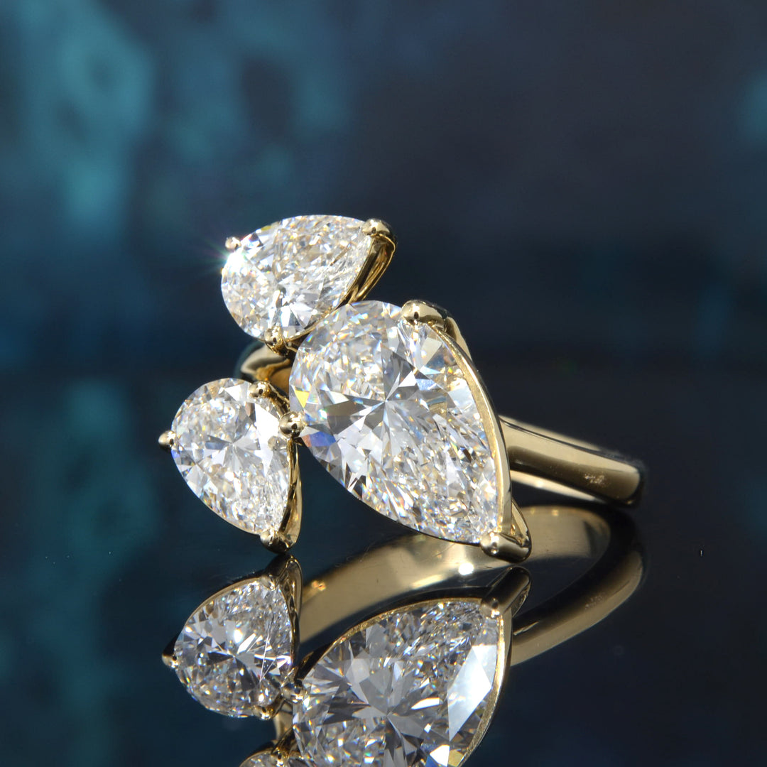 A yellow gold ring featuring three pear-cut diamonds in a cluster setting, displayed on a reflective dark blue surface.