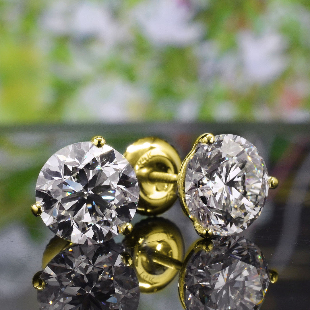 A pair of round-cut diamond stud earrings in yellow gold settings, shown on a reflective dark surface with a blurred green and white background.
