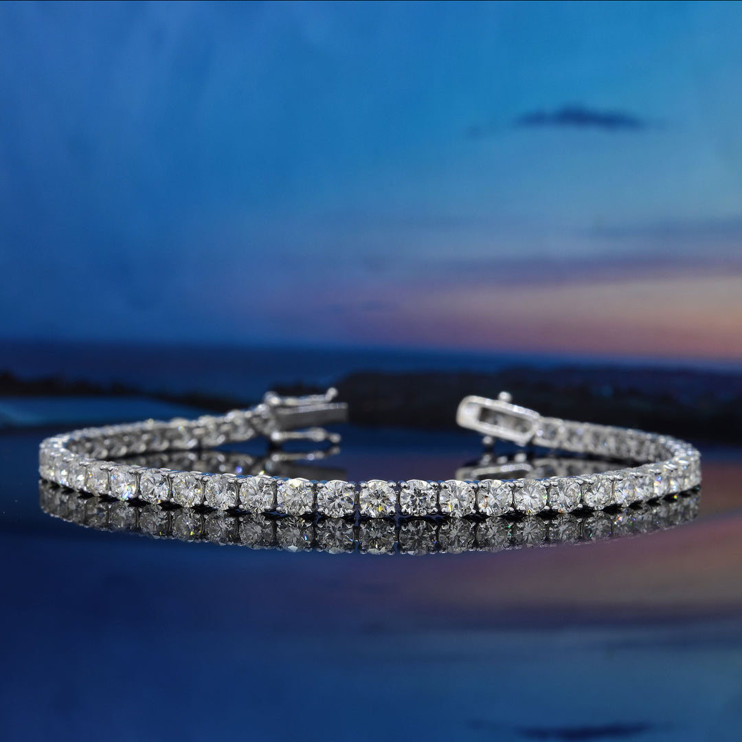 A silver tennis bracelet set with round clear stones, displayed on a reflective dark blue surface against a blurred blue sky and water background.