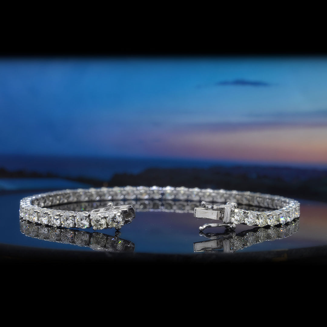 A diamond tennis bracelet in a silver-toned setting, displayed on a reflective dark surface with a blurred blue sky and ocean background.