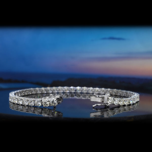 A diamond tennis bracelet in a silver-toned setting, displayed on a reflective dark surface with a blurred blue sky and ocean background.