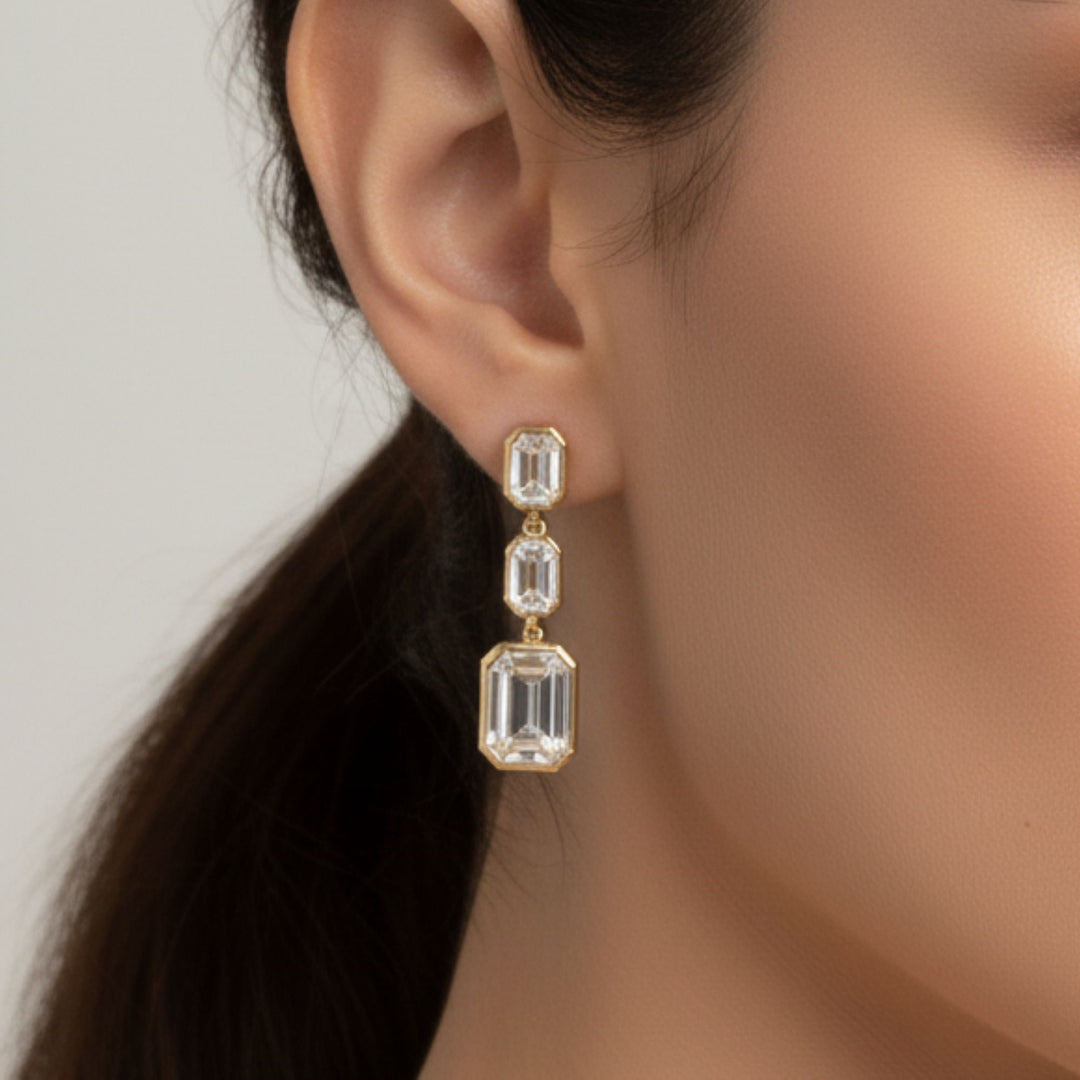 3 Emerald cut diamond dangling bezel set earrings in yellow gold worn on a lady's ear.