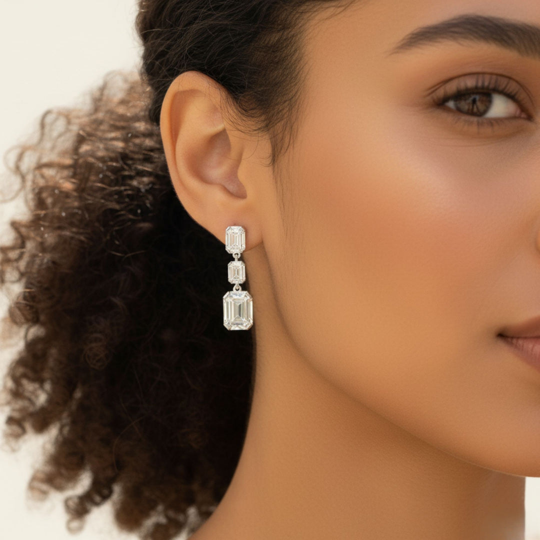 3 Emerald cut diamond dangling bezel set earrings in white gold worn by a lady model.