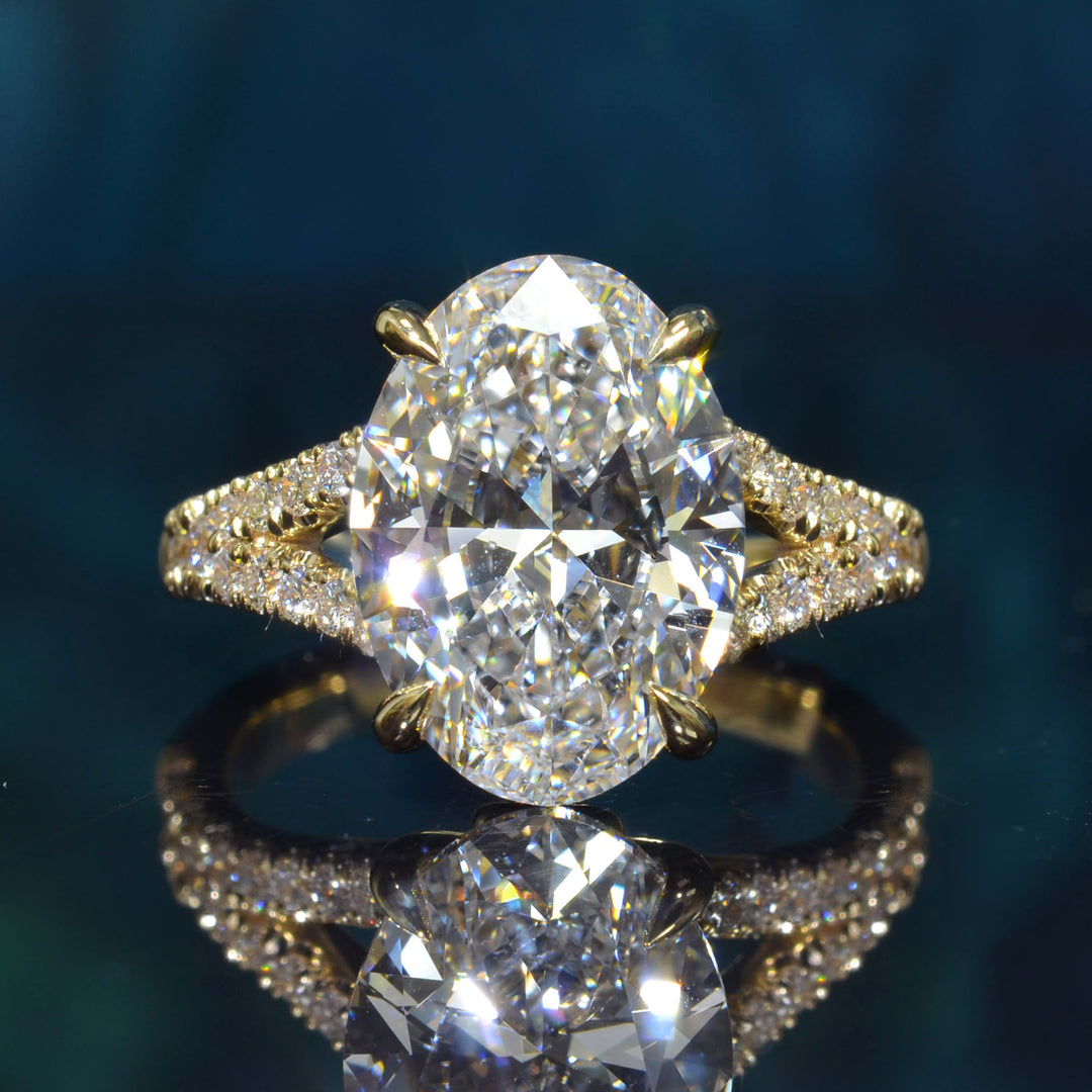 An oval-cut diamond engagement ring in yellow gold with a pavé band, displayed on a reflective dark teal surface.