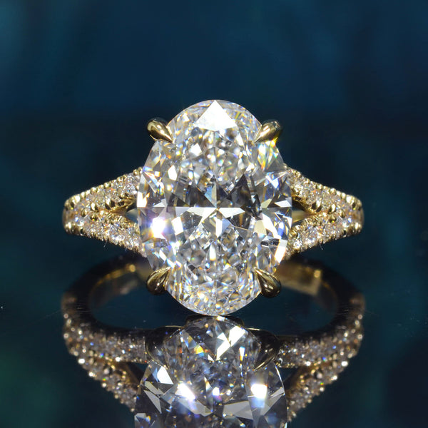 An oval-cut diamond engagement ring in yellow gold with a pavé band, displayed on a reflective dark teal surface.