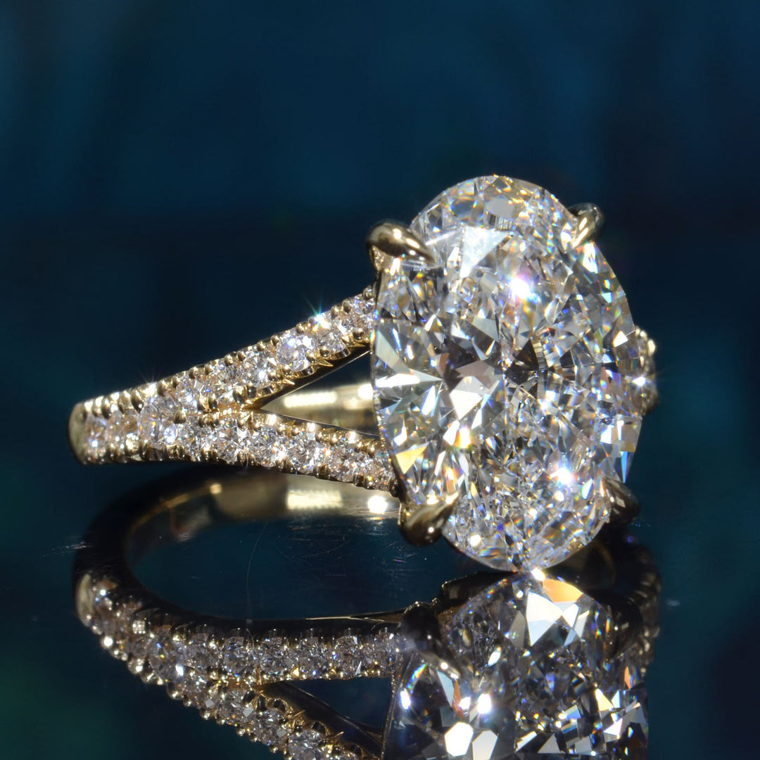 An oval-cut diamond engagement ring with a yellow gold band paved with small diamonds, shown on a reflective dark blue surface.