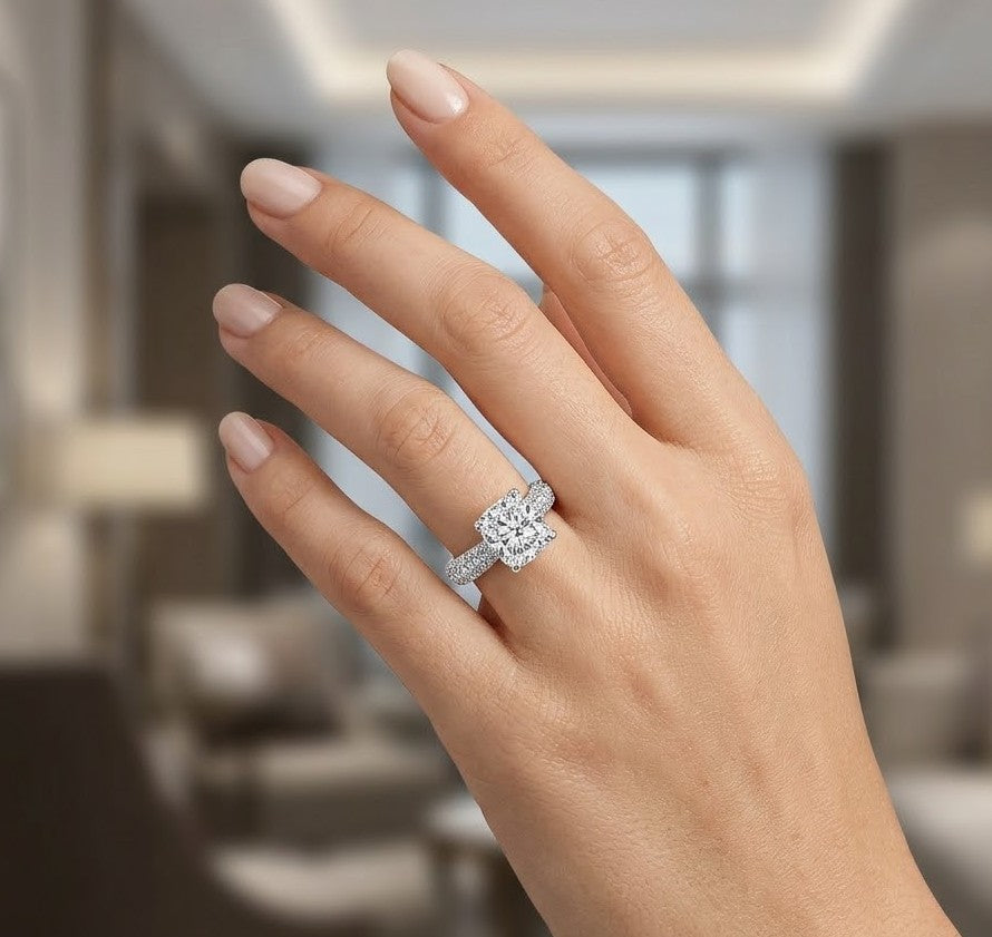 A close up of a hand wearing a diamond engagement ring with a cushion cut center stone and a pavé band, set against a softly blurred modern interior background