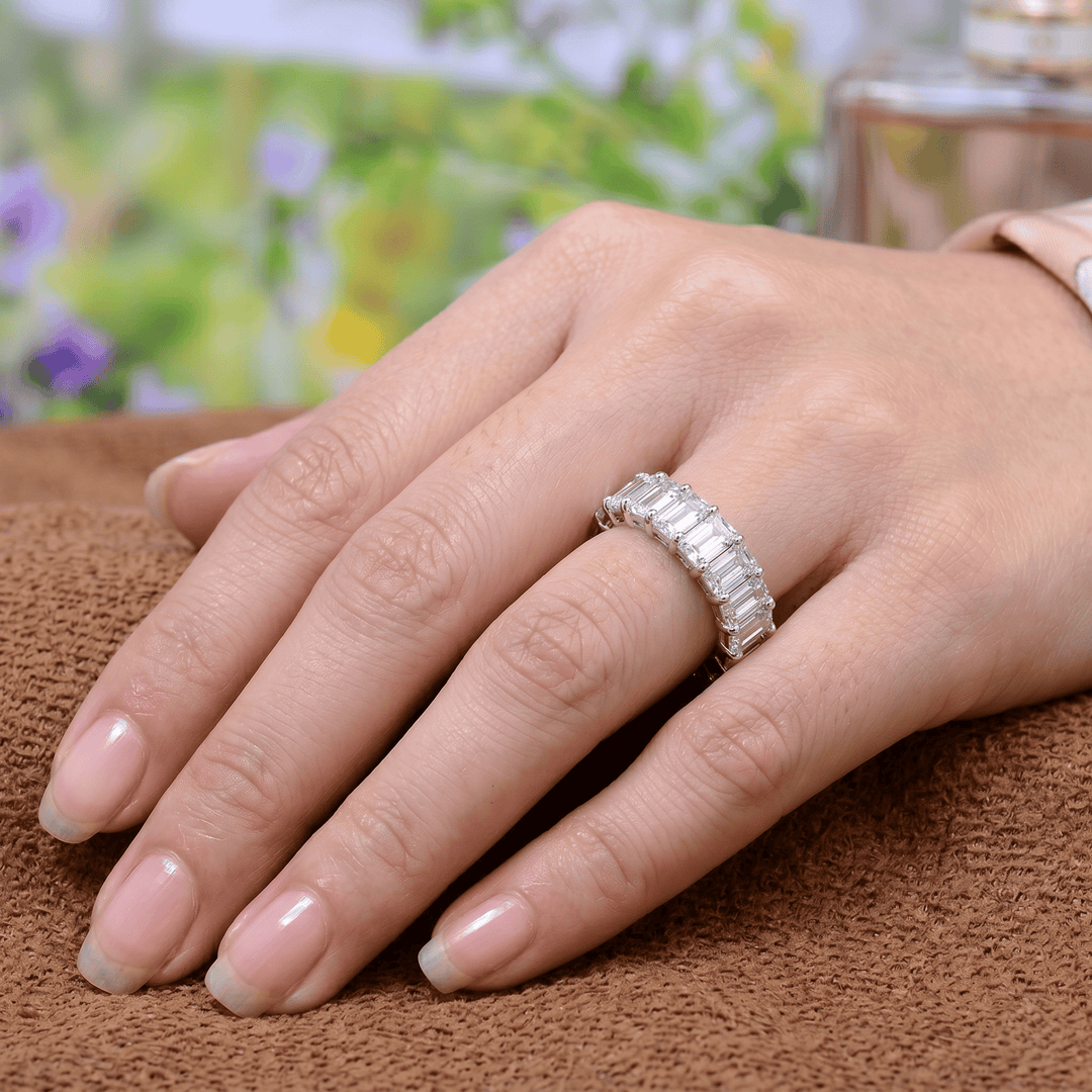 Hand wearing an eternity ring with emerald cut elongated diamonds on a brown surface with a blurred floral background