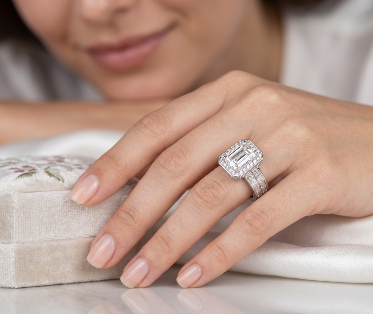 A close up of a woman's hand wearing an emerald cut diamond engagement ring with a diamond band, resting near a small embroidered jewelry box