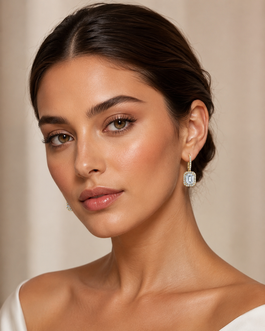 A close up portrait of a woman wearing a pair of rectangular drop earrings with clear diamonds in a silver toned setting, against a soft beige background