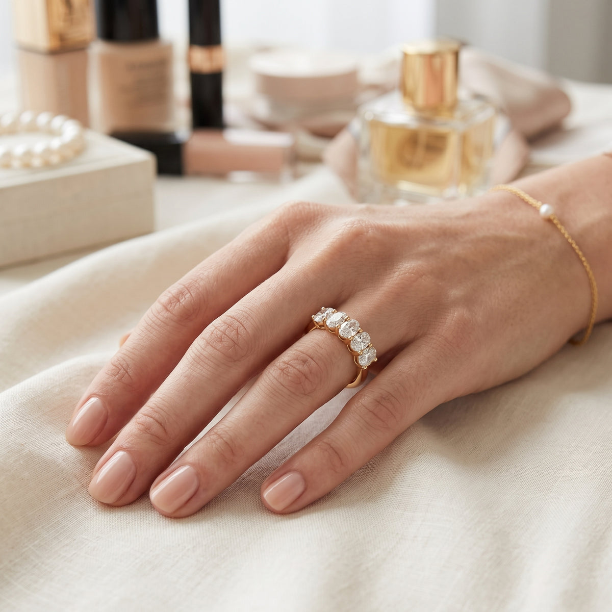 A close up lifestyle shot of a woman's hand wearing a gold band ring set with clear stones, resting on a soft beige fabric surface with cosmetic products blurred in the background